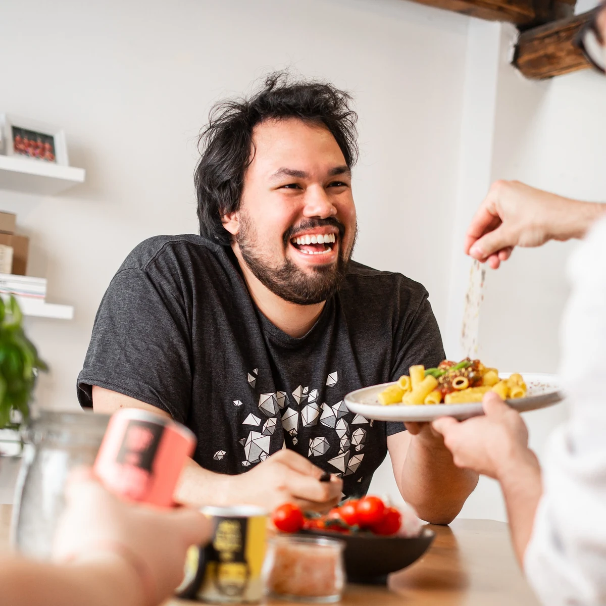 A smiling man at the table while a plate of pasta is seasoned in the foreground.